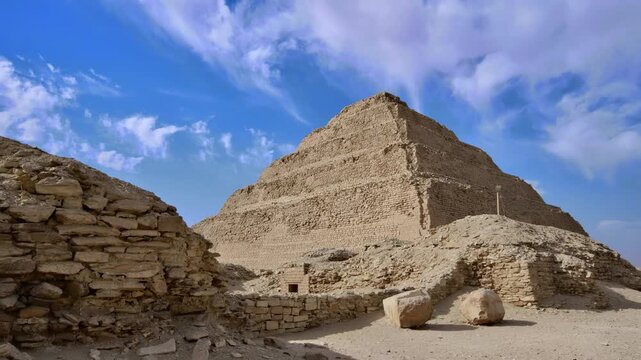 Step Pyramid of Djoser at Saqqara necropolis, Egypt, world's oldest pyramid with surrounding ancient ruins, stone blocks and arched entrance wall under blue sky.