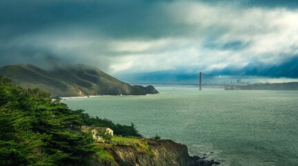 Naklejka premium Golden Gate Bridge partially visible through coastal fog with cliffs and ocean viewed from a scenic hillside in natural light.