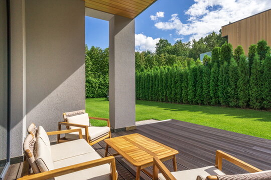 A small relaxation area features wooden chairs and a table. It overlooks a wide lawn with green grass and tall hedges. The sky is clear and sunny