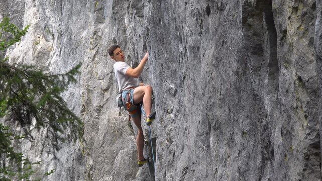 Strong male climber lead climbs a steep limestone wall, carefully moving between small holds on technical route. Balance, focus and athletic control during an outdoor sport climbing session in summer.