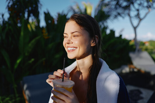 A smiling woman enjoys a refreshing drink outdoors in warm sun, wrapped in a light towel. Bright greenery and a serene setting convey relaxed lifestyle and summer leisure.