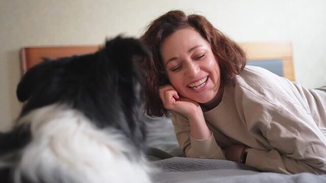 Smiling woman playing with cute puppy dog border collie on bed at home indoor. Owner girl stroking holding dog friend sitting on sofa in living room. Love for pets friendship support team concept
