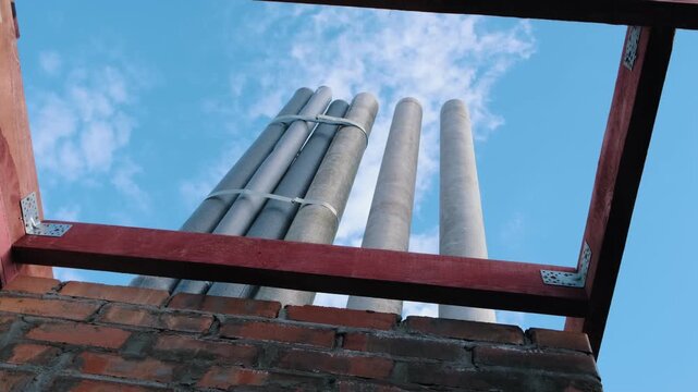 Low angle view of ventilation pipes rising above unfinished brick wall and wooden frame against blue sky, highlighting construction progress, building systems installation and residential development
