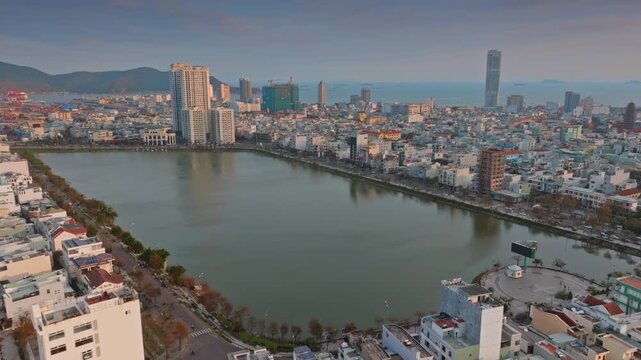 Aerial view of  Quy Nhon, Binh Dinh, Vietnam, with its modern skyline, a tranquil Ho Sinh Thai lake, and mountains in the background at sunset.