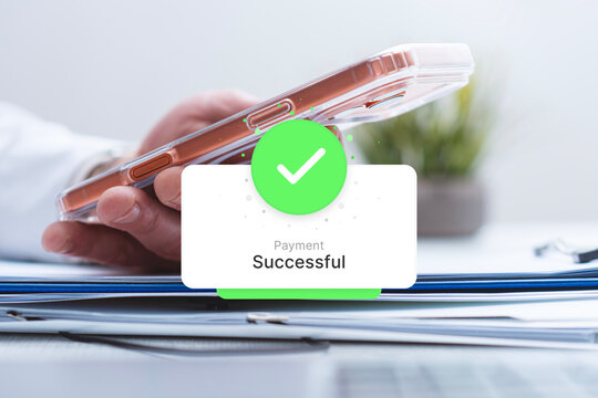 Hand holding a phone with a green payment successful notification pop up over a laptop. Concept for digital banking, online financial transactions, e-commerce checkout and business technology.