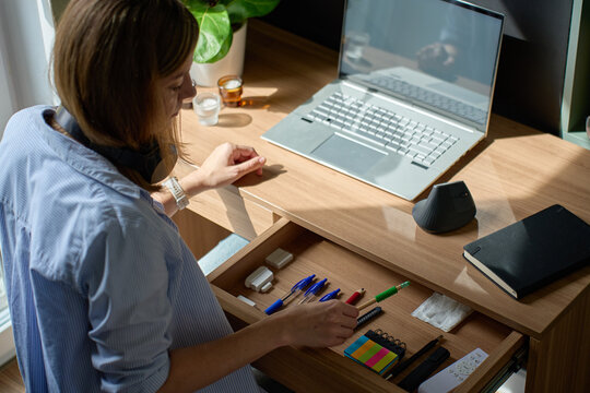 Woman arranging stationery in open desk drawer near laptop in home office