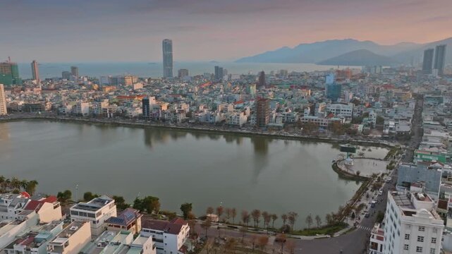 Aerial view of  Quy Nhon, Binh Dinh, Vietnam, with its modern skyline, a tranquil Ho Sinh Thai lake, and mountains in the background at sunset.