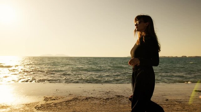 Young woman in black carrying roller skates along a sunlit wooden promenade by the sea. Golden sunset light and lens flare create cinematic atmosphere. Islands on horizon. Active coastal lifestyle 