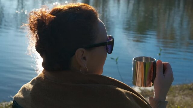 Woman enjoying a hot drink outdoors near a tranquil lake. Relaxed woman with red hair in sunglasses sitting by a lake, holding a steaming metal mug and enjoying the nature view during a sunny day
