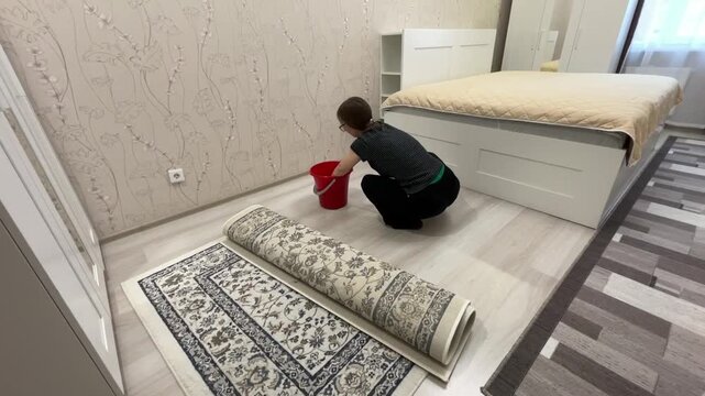 White woman scrubbing rug by bed with bucket and cloth, focused kneeling action, patterned runner partially rolled, bright modern bedroom, careful domestic routine and surface refresh