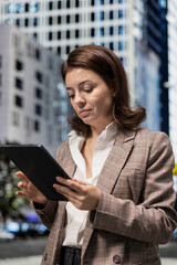 Fototapeta premium Businesswoman using tablet to browse online content, walking on city boulevard. Person using touchscreen device, relaxing by looking at social media posts on feed, urban cityscape background