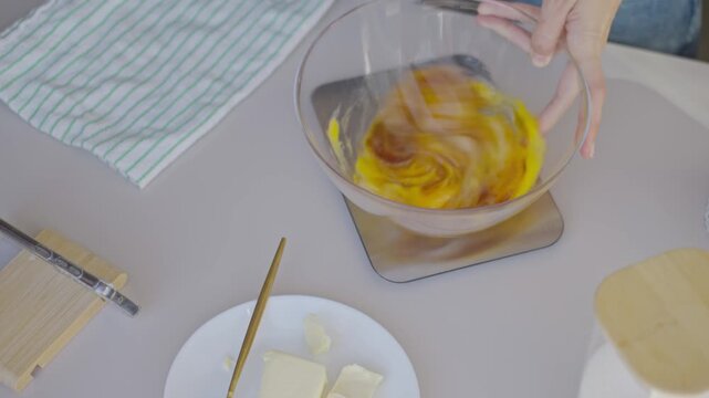 Woman whisks eggs and liquid ingredients in transparent bowl on kitchen table. Concept of baking and homemade cooking