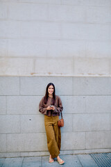 Fototapeta premium Young woman smiling, standing outdoors by a wall, wearing a brown cardigan and brown trousers, using a mobile phone