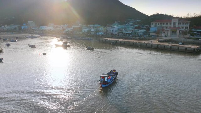 A fishing boat with several people on board travels through the water near a coastal town iof Hai Minh, Quy Nhon, Binh Dinh, Vietnam. The boat is leaving a wake as it moves.