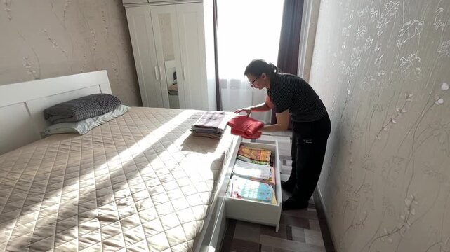 Sunlight highlights neatly stored bedding. Efficiently organizing linens with sunlight streaming through window. Woman carefully arranges soft towels and linens in wellorganized drawer