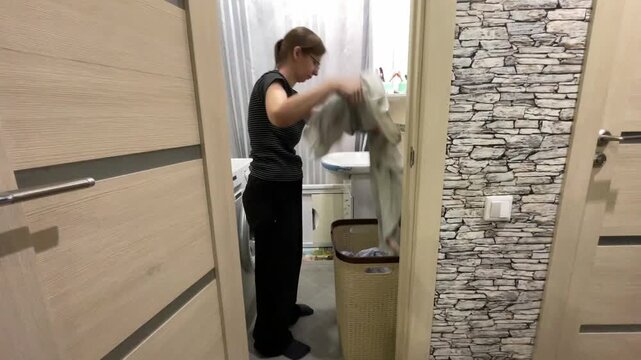 Compact bathroom scene, white woman sorts clothes into wicker hamper and tidies area near washer, methodical housekeeper organizing laundry with steady motions and neutral lighting