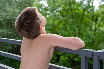 Obraz premium Young Shirtless boy standing on terrace, balcony, enjoying fresh air outdoors, summer day, peaceful Back view, green forest background, childhood moment, nature connection