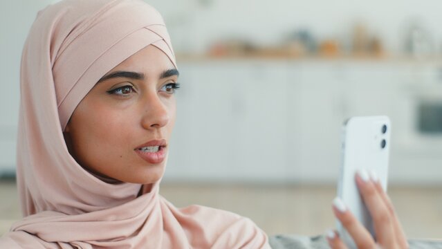 Smiling friendly Arabian Muslim woman in hijab talking video call indoors at home female Islamic girl young lady using mobile phone vlog blog smartphone chatting conference online virtual meeting