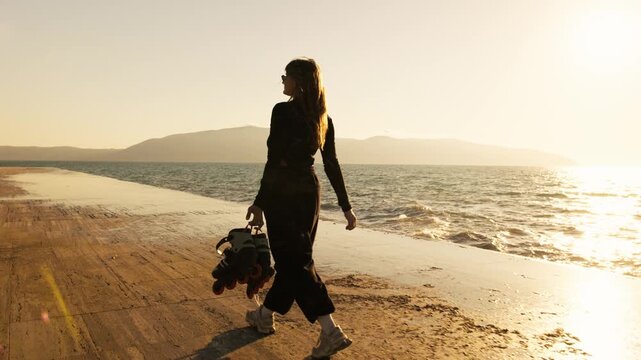 Rear view of woman in black carrying roller skates toward golden sunset on seafront. Sea reflection, mountain silhouettes, lens flare. Active summer freedom and outdoor lifestyle.
