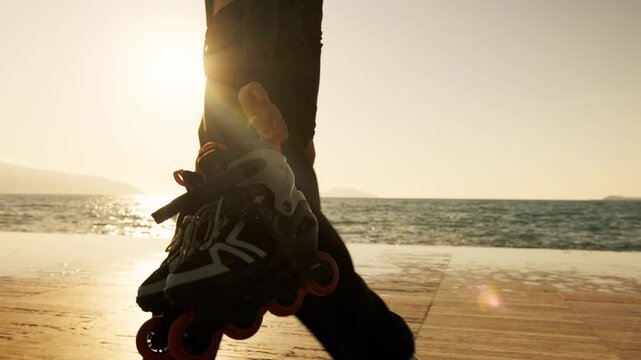Close-up of hand carrying orange-wheeled inline skates along golden sunset seafront. Silhouetted legs, sparkling sea, lens flare, mountain horizon. Active summer sport and freedom.
