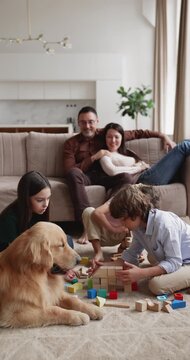 Vertical shot of large Caucasian family with golden retriever dog spending leisure time in living room, children together playing, building with wooden blocks while parents relaxing on couch at home