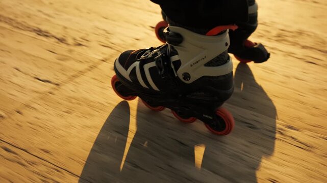 Extreme close-up of orange-wheeled inline skates gliding on golden wooden promenade at sunset. Sharp boot and lace detail, shadow beneath, motion blur. Active summer sport, speed and energy.
