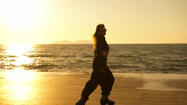 Full length silhouette of woman on roller skates glancing back at vivid golden sunset on seafront. Sparkling sea, islands, long shadow on glowing deck. Dynamic active summer freedom.
