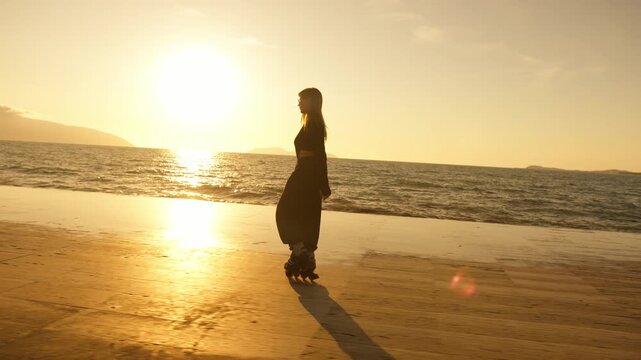 Cinematic silhouette of woman on roller skates gliding along golden sunset promenade by the sea. Long shadow, lens flare, mountain horizon. Freedom, active sport and summer lifestyle concept.
