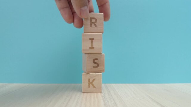 Close-up of a hand placing a wooden block with the letter R on top of a stack of blocks to complete the word RISK; the tower is shaking. Concept of business instability and risk management