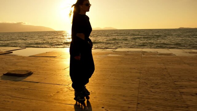 Full length silhouette of woman on roller skates skating toward camera against blazing golden sunset over sea. Dramatic halo, lens flare, mountain silhouettes. Active summer freedom and lifestyle.

