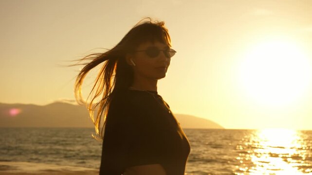 Close portrait of young woman skating at seafront in blazing golden sunset. Wind blows hair dramatically, warm backlight and lens flare. Sparkling sea, mountains. Freedom, active summer lifestyle.
