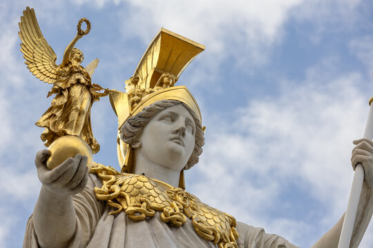 Low angle view of a white marble statue of the Greek goddess Athena wearing a golden helmet and holding a golden statuette of Nike against a cloudy blue sky.