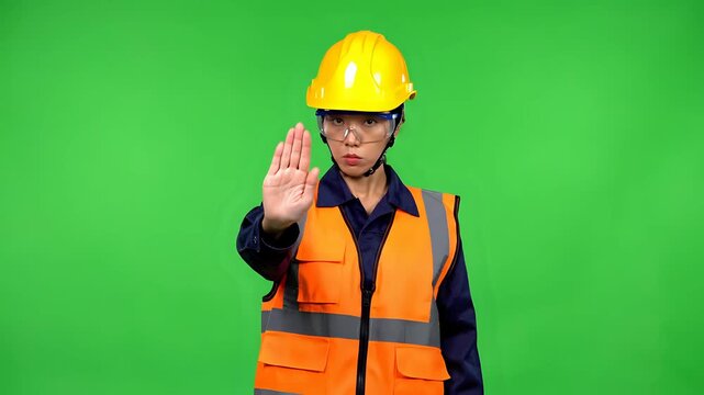 Construction worker in yellow hard hat and orange vest making stop gesture on green screen.