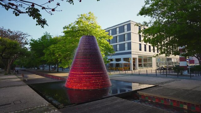 Large conical red and orange mosaic fountain with cascading water in urban square, green trees and modern building in background, slow motion, Lisbon