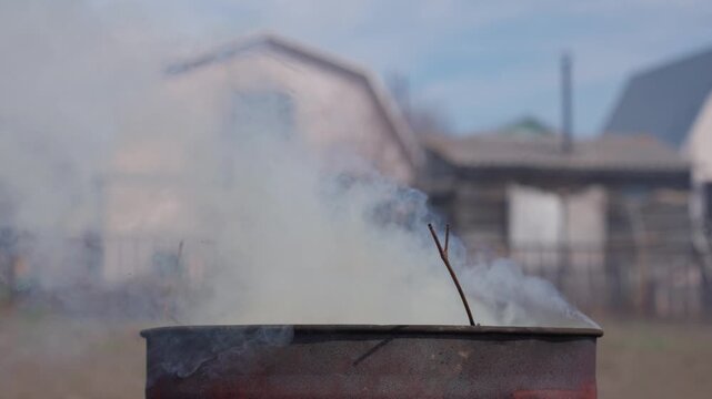 Rusted Burn Barrel Exhaling Pale Smoke, Blurred Houses In Background, Homesteader Performing Yard Cleanup, Thin Wisps Crossing Fence, Overcast Light Creating Contemplative Atmosphere And Muted Color