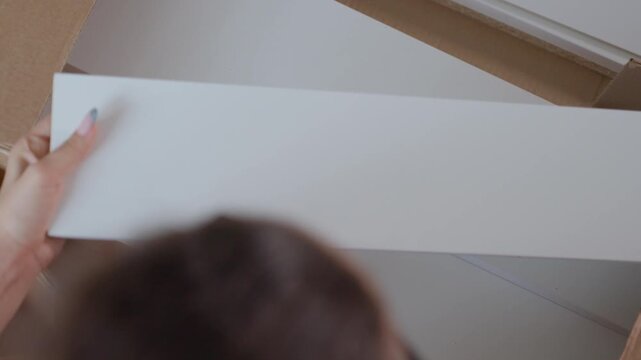 Woman Unpacking White Shelf Panels From Cardboard Box, Closeup Hands Slide Panel With Predrilled Holes, Careful Alignment For Flatpack Furniture Assembly, Domestic Living Interior Mood
