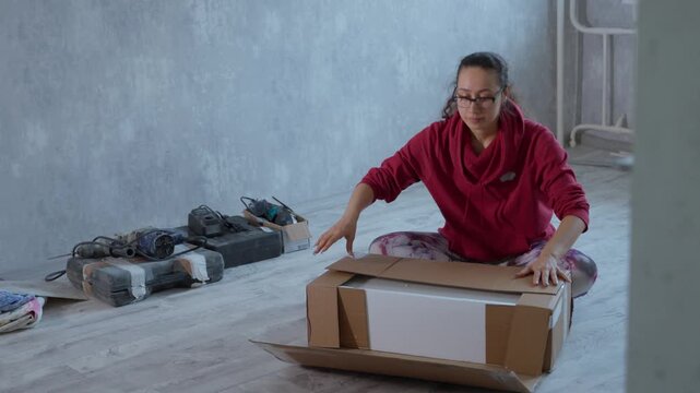 Woman Sorts Slowly. She Gently Places Box On Ground Among Tools And Unfolding Process. Woman Systematically Prepares Packing Box Close To Tools During Careful Unfolding And Arrangement
