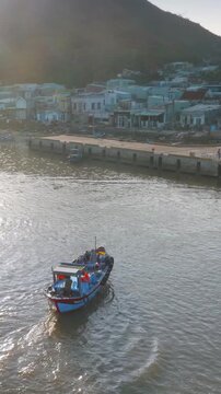 A fishing boat with several people on board travels through the water near a coastal town iof Hai Minh, Quy Nhon, Binh Dinh, Vietnam. The boat is leaving a wake as it moves.