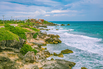 Scenic rocky coastline in Cancun, Mexico, with green vegetation and turquoise Caribbean waves crashing against rocks under a cloudy sky. © ZL Visuals