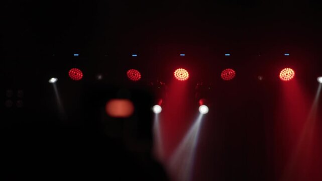 Lighting rig with red LED par lights and white moving head spotlight, warm glow on left with blurred crowd silhouette in foreground