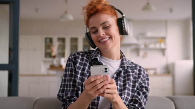 Happy smiling Caucasian woman dancing at home listening music in headphones relaxed female holding mobile phone using sound app online enjoying song audio carefree girl renter moving freely indoors