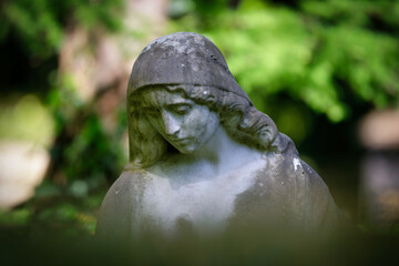 Fototapeta premium Close-up of a melancholy stone female statue with a downward gaze against a softly blurred green natural background