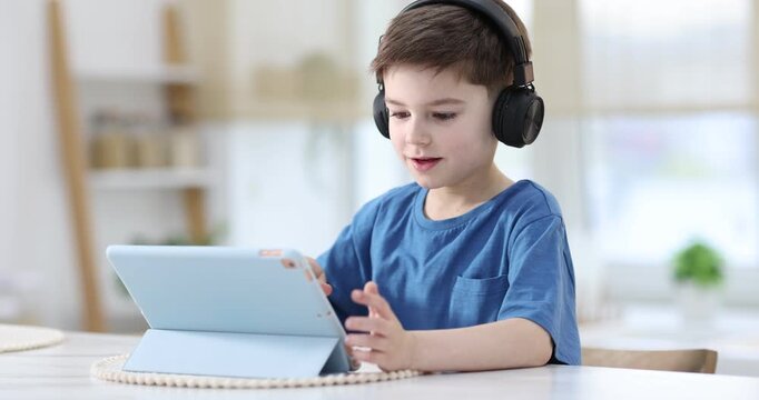 Little boy with headphones using modern tablet at white marble table indoors