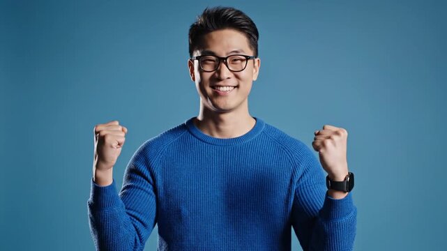 A young Asian man celebrating with fists clenched in a blue sweater on a blue background in a video