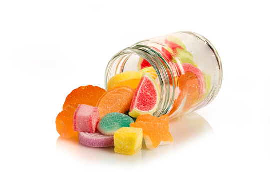Assorted vibrant sugar-coated fruit jelly candies in various shapes spilling out of glass jar. Sweet and delicious treat concept isolated on white background.