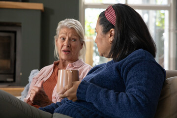 Naklejka premium Senior female friends chatting while holding ceramic mugs on living room sofa near fireplace
