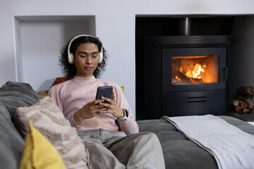 Naklejka premium Non-binary person lying on gray sofa at home, wearing white headset and checking phone