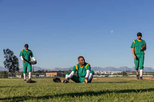 Diverse male teammates sitting and walking on outfield grass in green jerseys, tending gloves