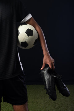 African man holding soccer ball and cleats wearing white-trim jersey on artificial turf, copy space