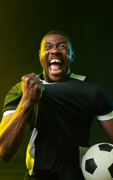Mid-adult African man shouting while gripping black jersey and holding soccer ball in studio